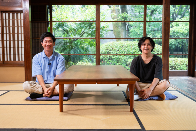 2人の男性が、机を挟んで和室に座ってこちらを向いている
